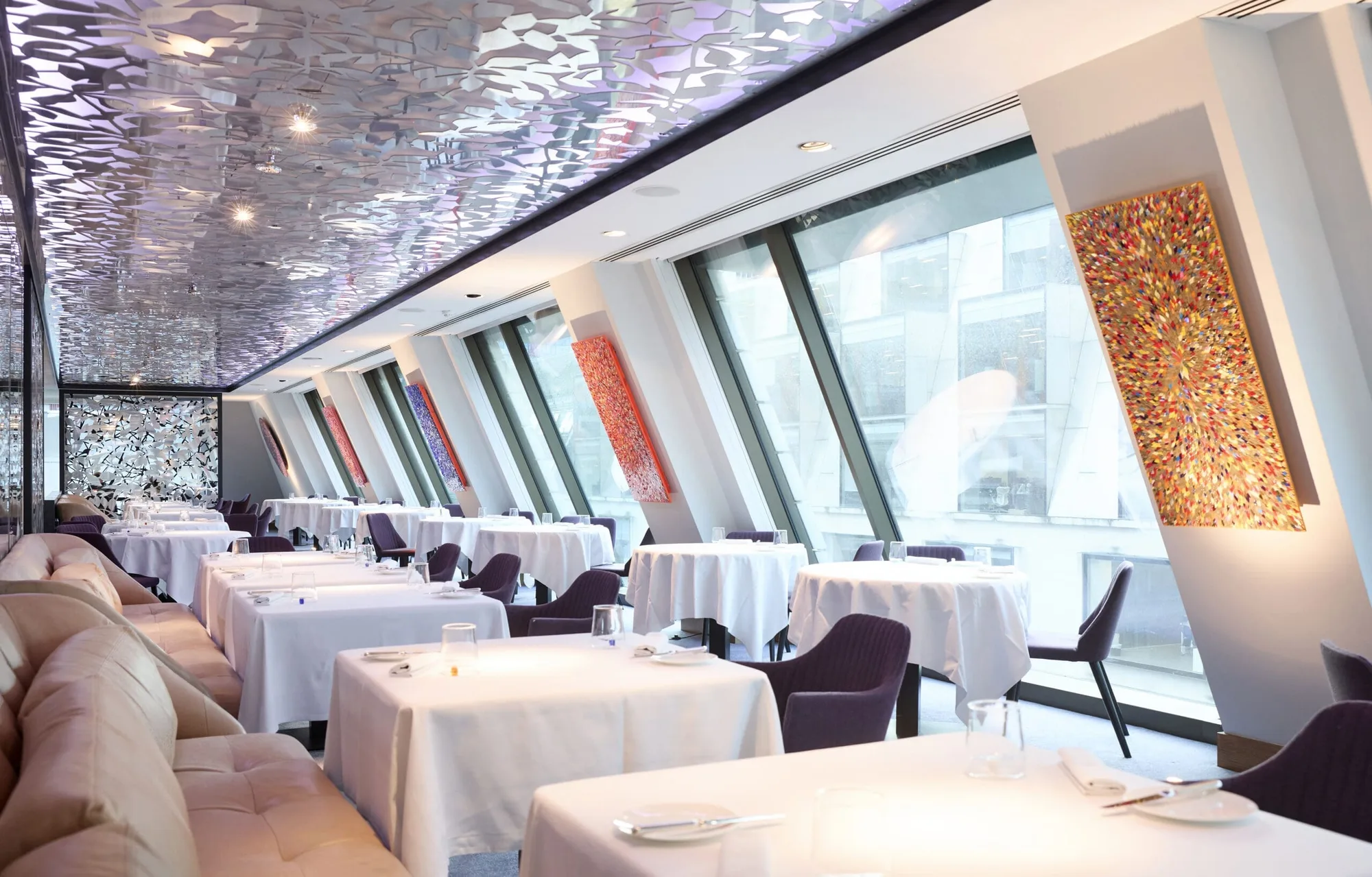 A modern, upmarket restaurant with large slanted windows, white tablecloths, purple chairs, abstract wall art, and a decorative reflective ceiling. Natural light fills the elegant dining area.