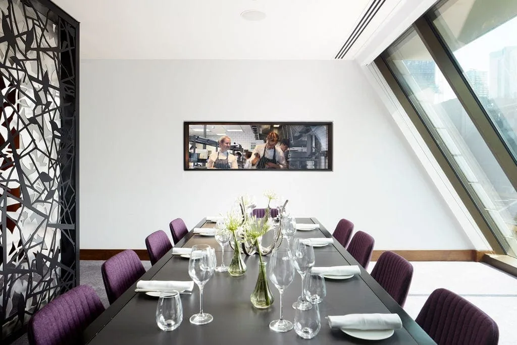 A modern dining room with a long black table set for eight, purple upholstered chairs, glass vases of flowers, and a wall photo of chefs cooking. Large slanted windows let in natural light.