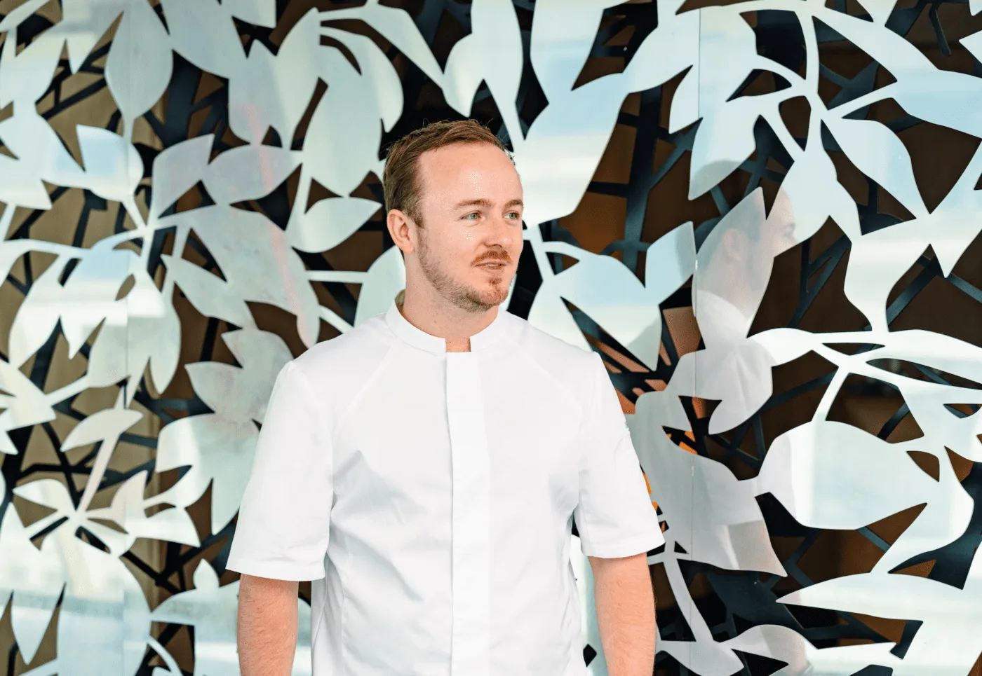 A man wearing a white chef’s jacket stands in front of a decorative wall with a leafy, abstract pattern, looking slightly to the side with a neutral expression.