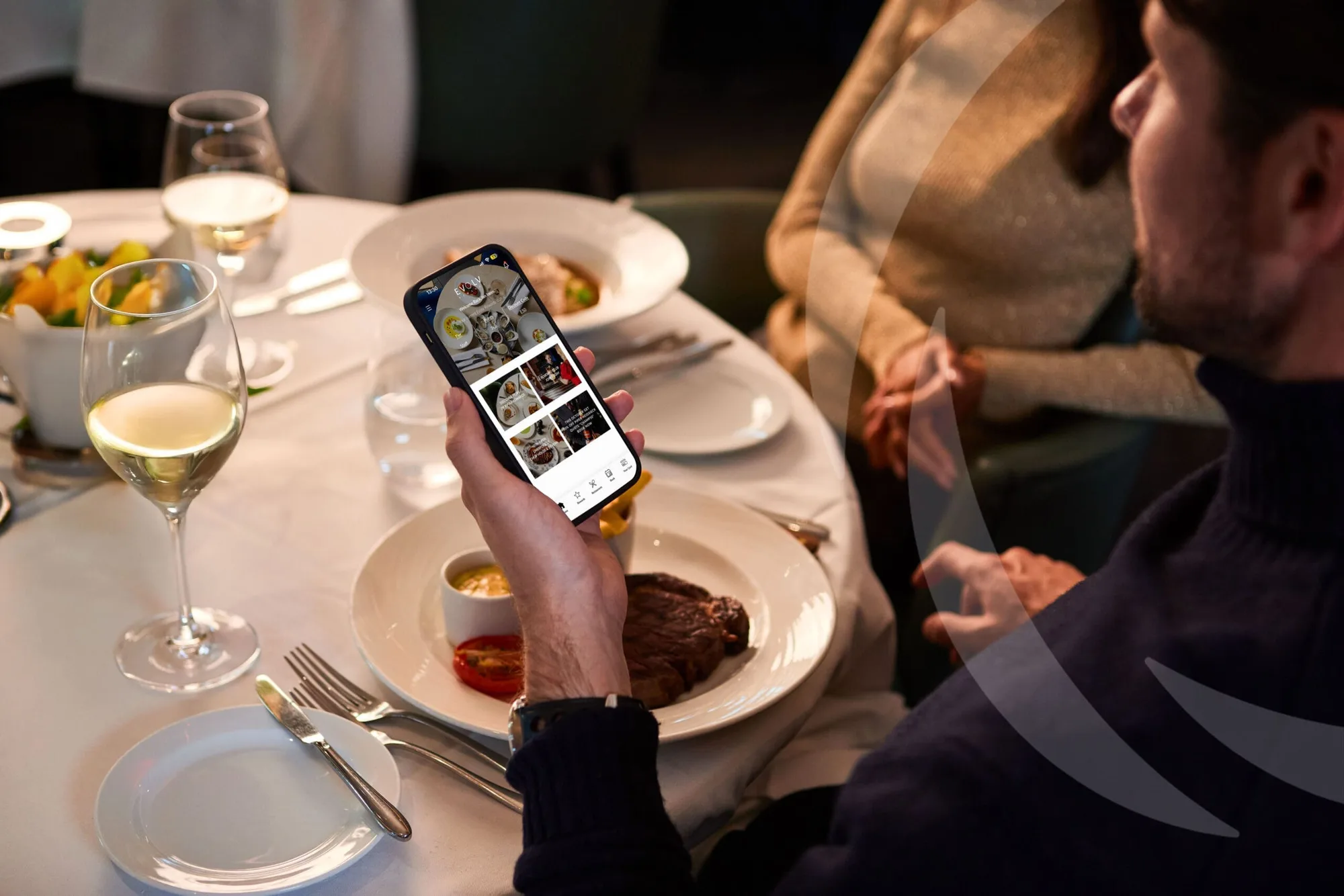 A man sits at a restaurant table with a plated steak meal, holding a mobile displaying a food delivery or menu app. A woman sits opposite him, and wine glasses and tableware are visible.