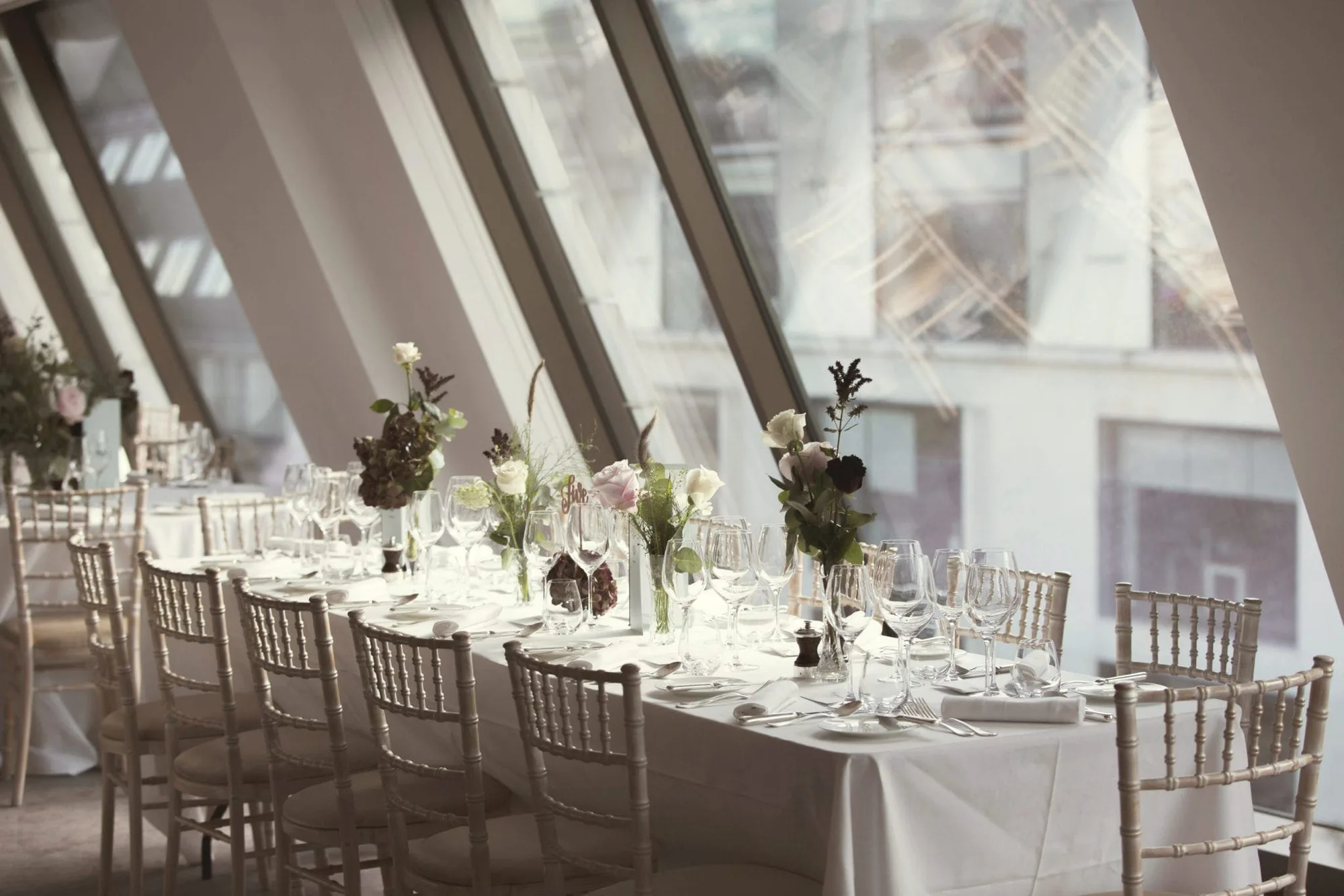 A long, elegant dining table set with white linen, glassware, plates, and floral centrepieces stands beside large slanted windows, letting in natural light. Cream chairs are neatly arranged along both sides of the table.