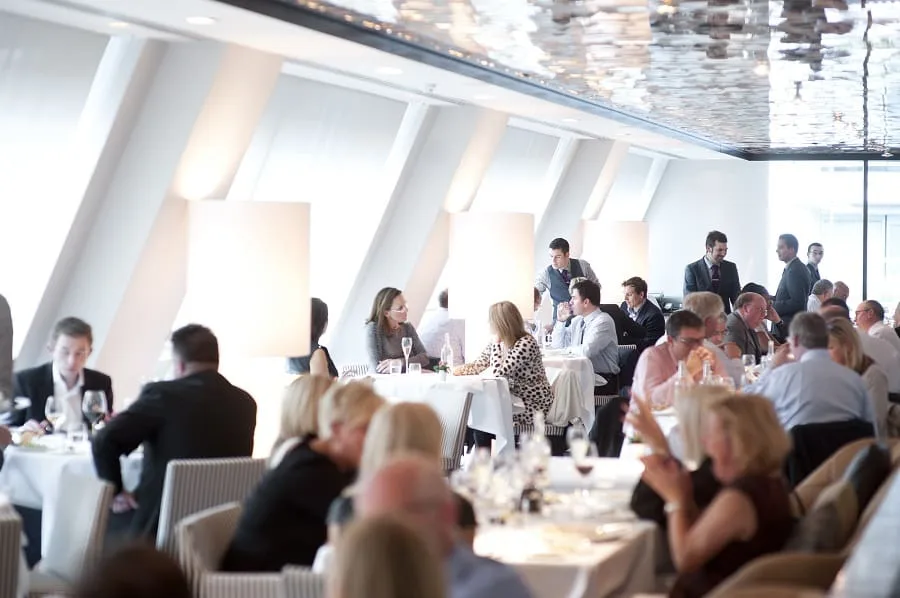 A busy, modern restaurant filled with people dining at white-clothed tables, engaged in conversation. Soft natural light streams through large windows, creating a bright, lively atmosphere.