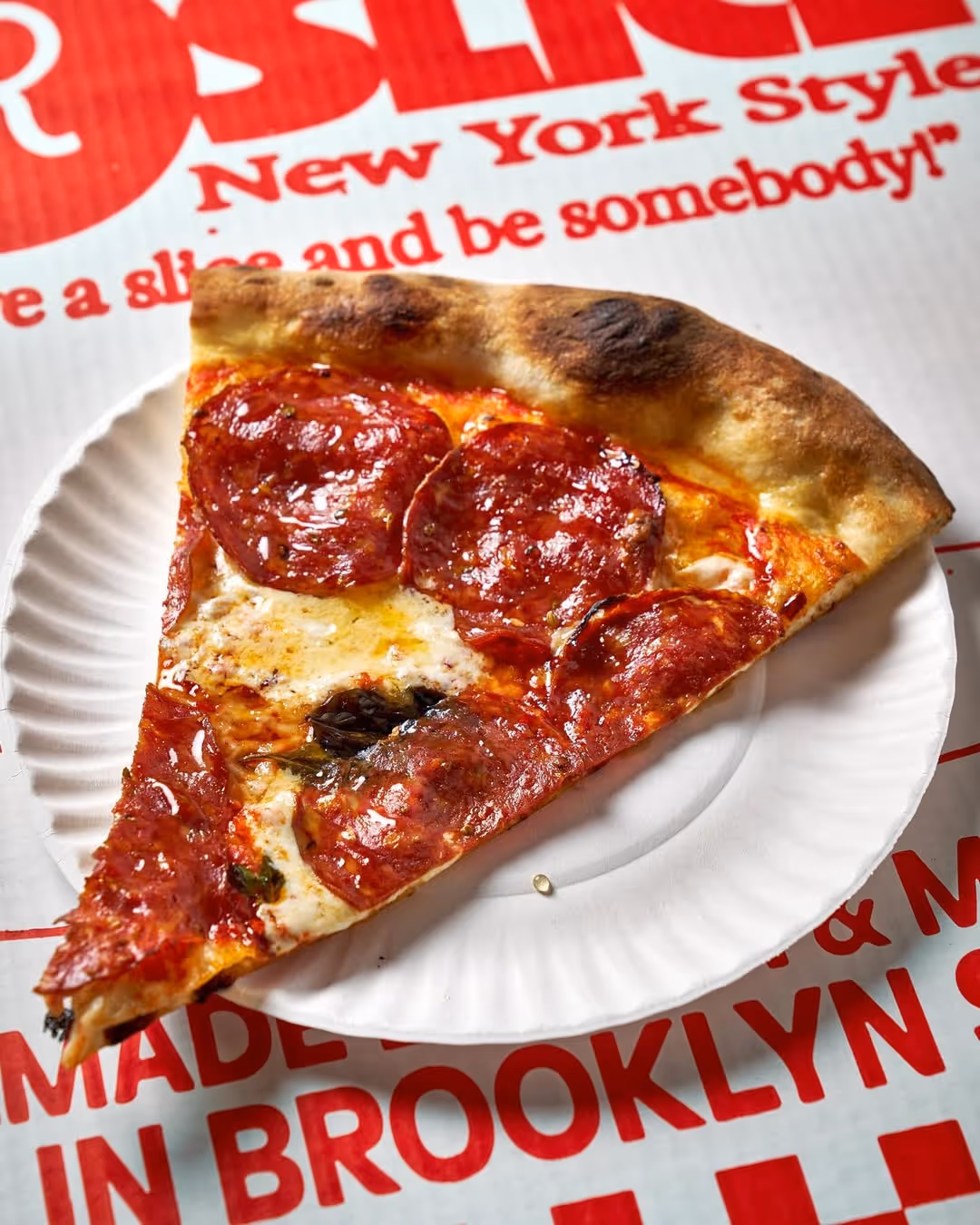 A crispy slice of pepperoni pizza on a paper plate, resting on a pizza box from Roberta’s Pizza restaurant, the famous New York-style pizzeria in Brooklyn.