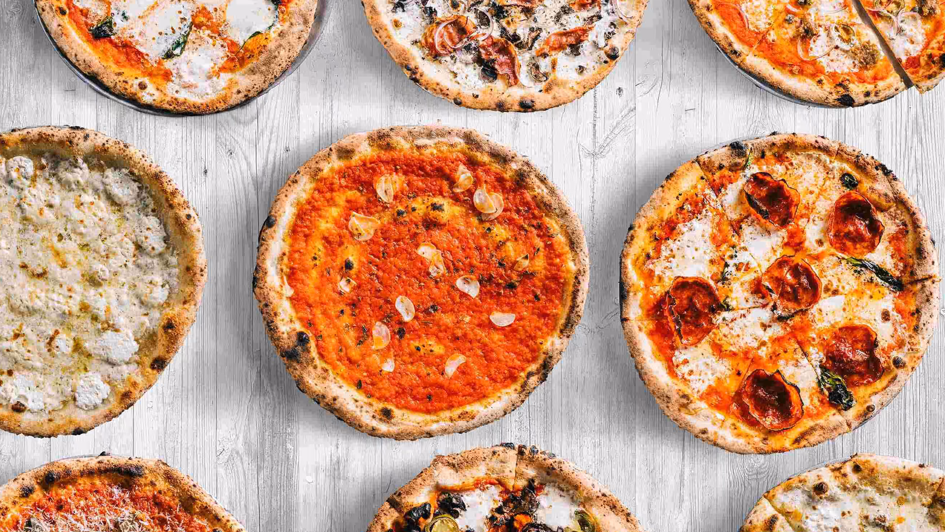 Top-down view of various artisanal pizzas, including Margherita and white pies, on a light wood table at Roberta’s Pizza restaurant.